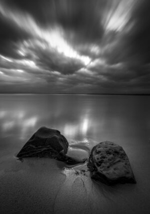 A black and white photograph shows a short sandy beach with two large rocks. The sun is setting thorugh clouds and reflecting on the water.