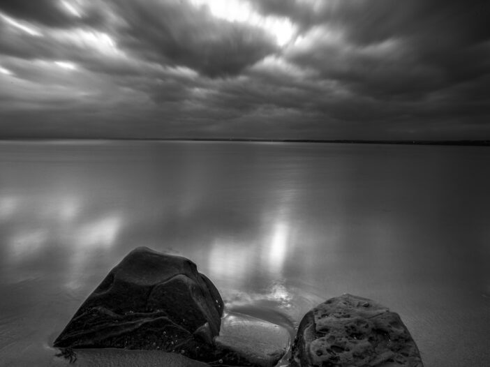 A black and white photograph shows a short sandy beach with two large rocks. The sun is setting thorugh clouds and reflecting on the water.