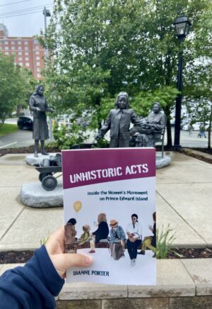 A copy of Unhistoric Acts is held in front of a statue to women volunteers of World War Two.