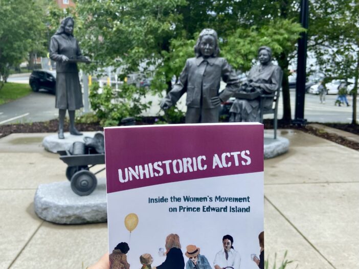A copy of Unhistoric Acts is held in front of a statue to women volunteers of World War Two.