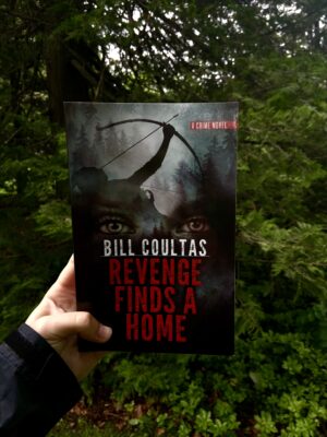 A copy of Revenge Finds a Home is held in front of a dark and grim forest.