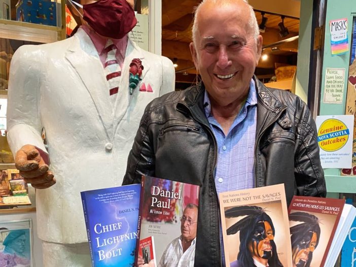 A smiling man stands next to a chainsaw-carved statue of a former prime minister. The man is holding a handfuls of books